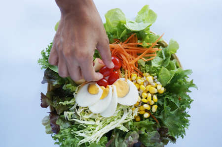 A photo of fresh vegetables prepare for salad, corn, tomatoes, apple, cabbage, lettuce with boiled eggs and hand pick up a tomato isolated on white background, close upの写真素材