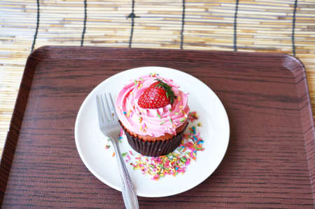 A photo of colorful cream strawberry cupcake decorated with sugar topping on white plate on wooden tray on rattan curtain background, close upの写真素材