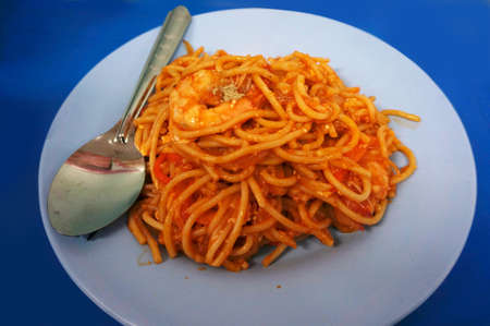 Thai Italian fusion food mix, Stir fried spaghetti pasta with ketchup, shrimps and tomatoes on white plate on blue backgroundの写真素材
