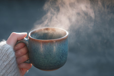 Female hand holding a cup of hot coffee with steam on a dark backgroundの素材