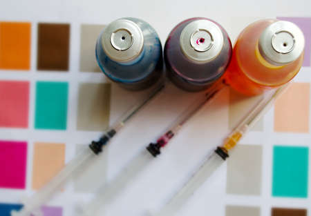Bottle of ink printer has three colors. Colors is red, yellow and blue with syringe on colored paper.の写真素材