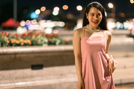 A confident young woman in a pink dress stands outdoors at night, radiating elegance and charm against a backdrop of glowing bokeh lights and floral arrangements.の写真素材