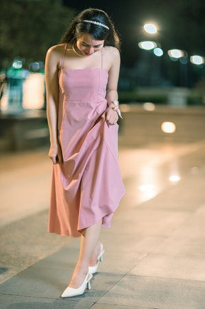 A stylish young woman in a beautiful pink dress walks gracefully along a well-lit urban street at night, exuding elegance and joy amidst a serene atmosphere.の写真素材