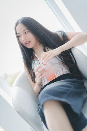 A young woman sits comfortably in a modern indoor space, sipping a refreshing pink drink. Her relaxed expression conveys joy and a carefree lifestyle, perfect for summer vibes.の写真素材
