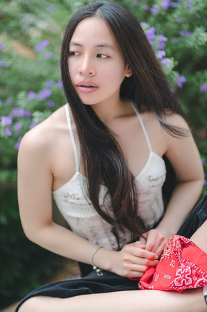 A young woman with long hair is sitting in a beautiful natural setting filled with flowers. She...
