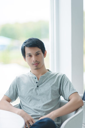 A young man in casual gray clothing sits thoughtfully by a window, capturing a moment of peace and contemplation in a modern indoor space filled with natural light.の写真素材