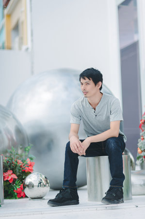 A young man sits thoughtfully on a metallic stool in a contemporary indoor space, surrounded by sleek sculptures and vibrant flowers, creating an artistic atmosphere.の写真素材