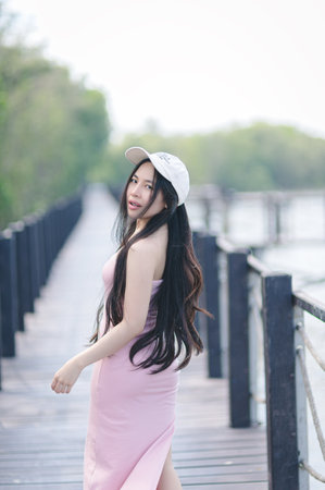 A joyful young woman in a pink dress stands on a scenic wooden pathway, surrounded by lush greenery and a tranquil water body, embodying summer leisure and beauty.の写真素材