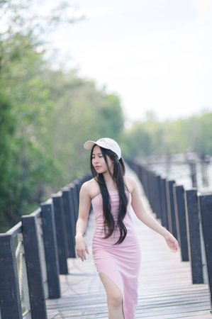 A young woman in a stylish pink dress walks gracefully on a wooden pathway, surrounded by lush greenery. The bright sunlight enhances the serene atmosphere.の写真素材