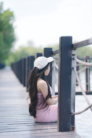 A young woman sits peacefully on a wooden dock by the water, enjoying a serene moment in nature. The soft sunlight enhances the calm atmosphere.の写真素材