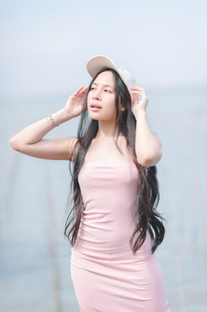 A young woman confidently poses by a tranquil lake, wearing a stylish pink dress and a cap. Her long hair flows gracefully as she enjoys the serene summer day.の写真素材