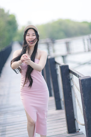A joyful woman in a pink dress smiles playfully on a scenic wooden pathway surrounded by lush greenery. Perfect for lifestyle and fashion themes.の写真素材