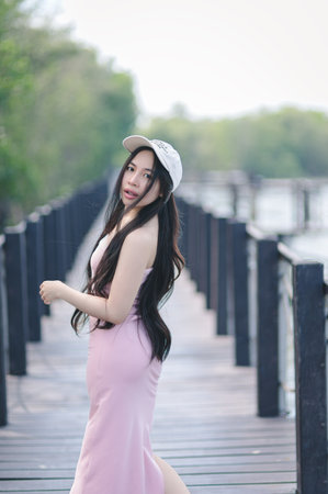 A serene portrait of a young woman in a pink dress walking gracefully along a boardwalk by the water. The image captures a tranquil outdoor setting.の写真素材