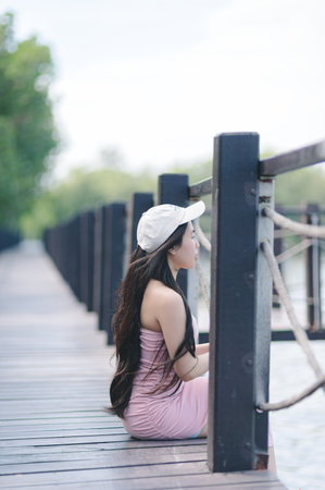A young woman relaxes by the water's edge, sitting on a dock. She wears a white cap and a stylish dress, embodying tranquility amid natureの写真素材
