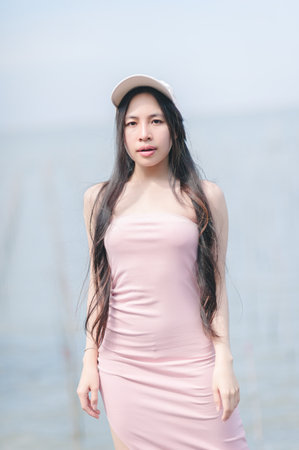 A young woman poses confidently in a pink dress and cap by the water's edge, exuding serene beauty and youthful charm in a natural outdoor setting.の写真素材