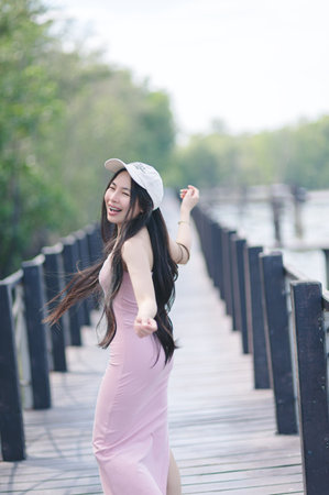 A joyful young woman in a pink dress dances along a wooden walkway surrounded by lush greenery. Her carefree motion and bright smile embody a spirit of happiness and freedom.の写真素材
