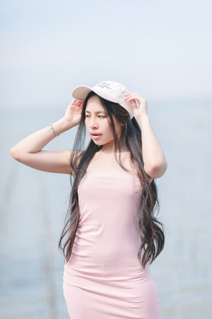A young woman showcases her style in a pink dress and hat by the water. With long hair flowing naturally, she embodies beauty and serenity in a vibrant outdoor setting.の写真素材