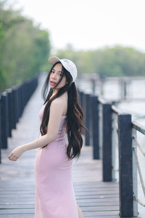 A young woman in a pink dress walks gracefully along a scenic wooden dock. The image captures her long hair flowing in the gentle breeze, showcasing a moment of peace and natural beauty amidst a serene landscape.の写真素材