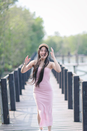 A cheerful young woman in a pink dress joyfully walks along a wooden walkway surrounded by lush greenery. Her long hair flows in the breeze, embodying freedom and happiness.の写真素材