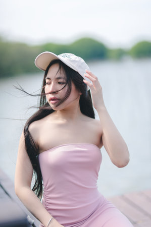 A young woman in a pink dress and cap poses gracefully by the water, embodying a sense of tranquility and summer vibes amidst the serene natural backdrop.の写真素材