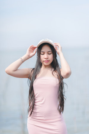 A young woman dressed in a pink dress captures a serene moment by the water. Her long hair flows gently as she enjoys the sunny day, radiating calmness.の写真素材