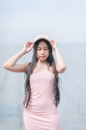 A young woman in a stylish pink dress and hat enjoys a serene moment by a tranquil lake, embodying beauty and calmness in a natural setting.の写真素材