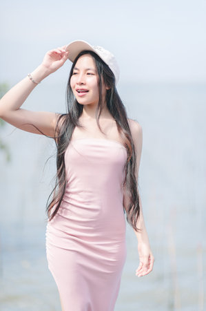 A young woman wearing a pink dress and a cap enjoys a sunny day by the water. Her long hair flows gracefully as she smiles, embodying the essence of joy and relaxation in a beautiful outdoor setting.の写真素材