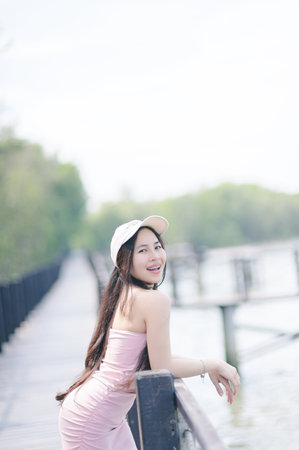 A cheerful young woman in a stylish dress smiles while standing on a boardwalk by the water. The serene environment showcases nature's beauty, perfect for leisure and relaxation.の写真素材