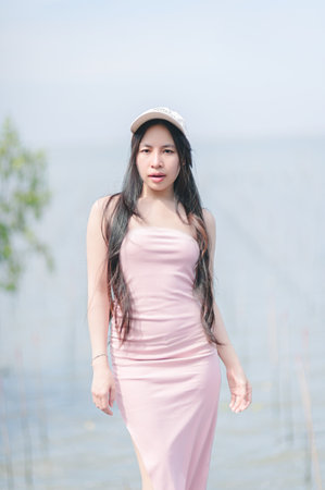 A young woman is captured standing gracefully by a tranquil lake, wearing a lovely pink dress and a cap. The serene environment reflects her beauty and confidence.の写真素材