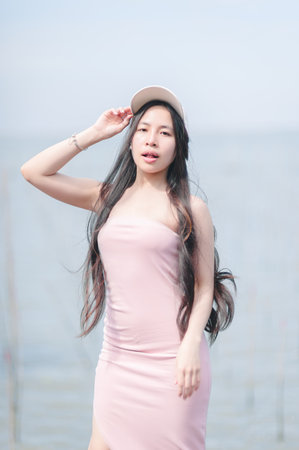 A young woman poses confidently in a pink dress near calm water, embracing the serene surroundings. Her stylish casual look and relaxed expression capture the essence of summer beauty.の写真素材