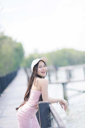 A cheerful young woman poses gracefully by a scenic river, wearing a stylish pink dress and a cap. Embrace the beauty of nature and joyful moments.の写真素材