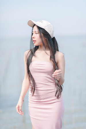 A young woman poses gracefully by the water's edge, wearing a pink dress and a cap. The serene setting captures summer vibes and natural beauty.の写真素材