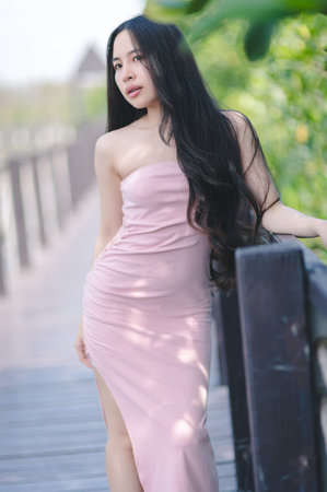A beautiful woman modeling an elegant pink dress, standing gracefully on a wooden walkway. Surrounded by lush greenery, her long hair flows gently in the breeze, evoking a serene summer vibe.の写真素材