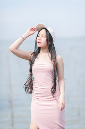 A young woman confidently poses in a pink dress by the water, radiating elegance and tranquility under the bright sunlight. Her casual hat adds a stylish touch.の写真素材