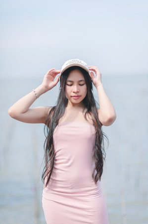 A young woman poses gracefully by the water, wearing a pink dress and a stylish hat. The soft colors and serene environment create a peaceful summer vibe.の写真素材
