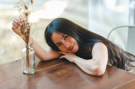 A serene image of a young woman resting her head on her hand while surrounded by dried flowers in a glass vase, creating a tranquil atmosphere in a bright indoor setting.の写真素材
