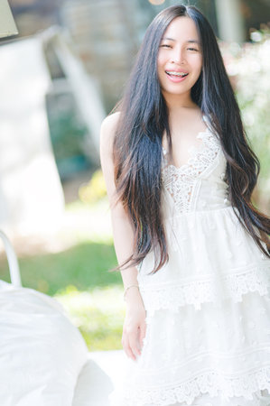 A joyful young woman with long hair smiles while wearing a white summer dress outdoors. The bright sunlight highlights her natural beauty in a serene setting.の写真素材