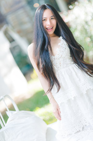 A young woman with long hair smiles brightly while wearing a white dress in a sunny outdoor setting, surrounded by green vegetation, exuding joy and elegance.の写真素材