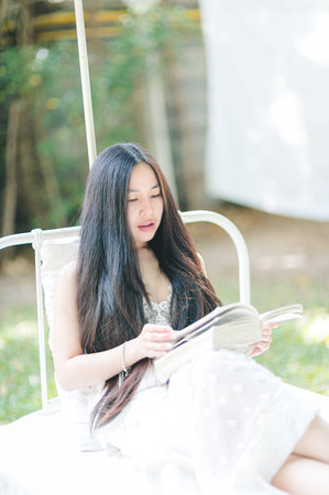A young woman enjoys a peaceful moment reading a book outdoors, surrounded by nature. Her long hair cascades down as she relaxes in a bright, tranquil environment.の写真素材