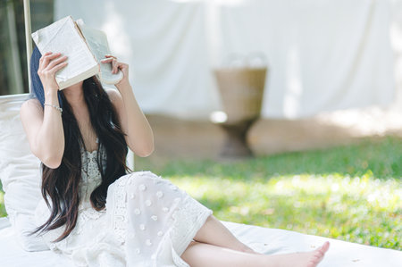 A woman sits peacefully outdoors, holding a book in front of her face, enjoying a tranquil moment in nature. The setting offers a serene atmosphere filled with soft light and greenery.の写真素材