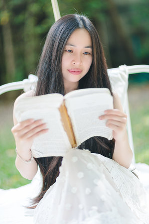 Serene young woman sits outdoors, reading a book in a soft white dress. Surrounded by nature and sunlight, she embodies tranquility and contemplation, enjoying a peaceful moment.の写真素材