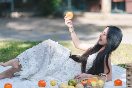 A young woman relaxes on a sunny day, surrounded by fresh fruits. She holds an orange, enjoying nature and embodying joy and vibrancy in her outdoor setting.の写真素材