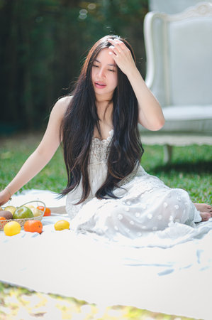 A serene young woman sits on the grass, surrounded by fresh fruits, enjoying the warmth of the sun. She embodies relaxation and natural beauty in a tranquil outdoor setting.の写真素材