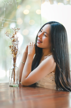 A joyful young woman smiles while sitting at a table with a simple flower arrangement. The soft bokeh background adds a warm and inviting atmosphere, highlighting her natural beauty and relaxed demeanor.の写真素材