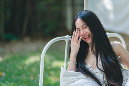 A joyful woman with long hair is sitting outdoors in a serene setting, enjoying the sunlight and a moment of relaxation. Her radiant smile conveys happiness and tranquility.の写真素材