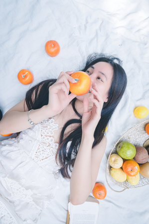 A serene image of a young woman enjoying a fresh orange while surrounded by vibrant fruits on a soft blanket. The scene captures a joyful and healthy lifestyle.の写真素材
