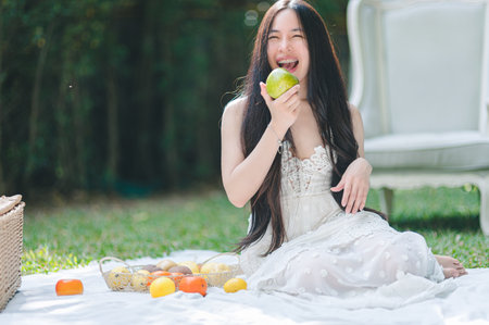 A joyful young woman sits on a blanket in a sunny garden, playfully tasting fresh fruits. The scene captures the beauty of nature and the essence of summer.の写真素材