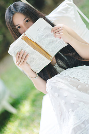 A young woman sits calmly outdoors, holding an old book. The sunlight filters through green surroundings, creating a peaceful and idyllic atmosphere for reading.の写真素材