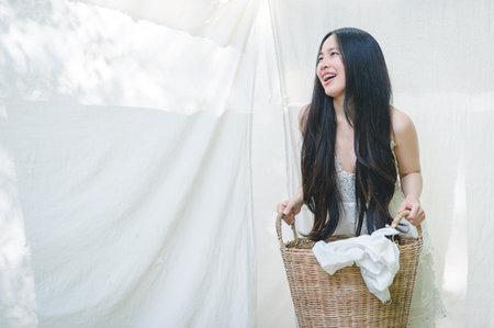 A young woman with long black hair smiles radiantly while holding a wicker basket filled with laundry. The setting features soft natural light, enhancing the cheerful ambiance.の写真素材