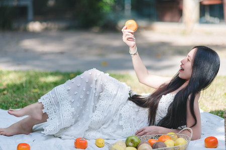 A joyful woman in a beautiful white dress lays on the grass, enjoying fresh fruit in a serene garden, capturing the essence of relaxation and nature.の写真素材